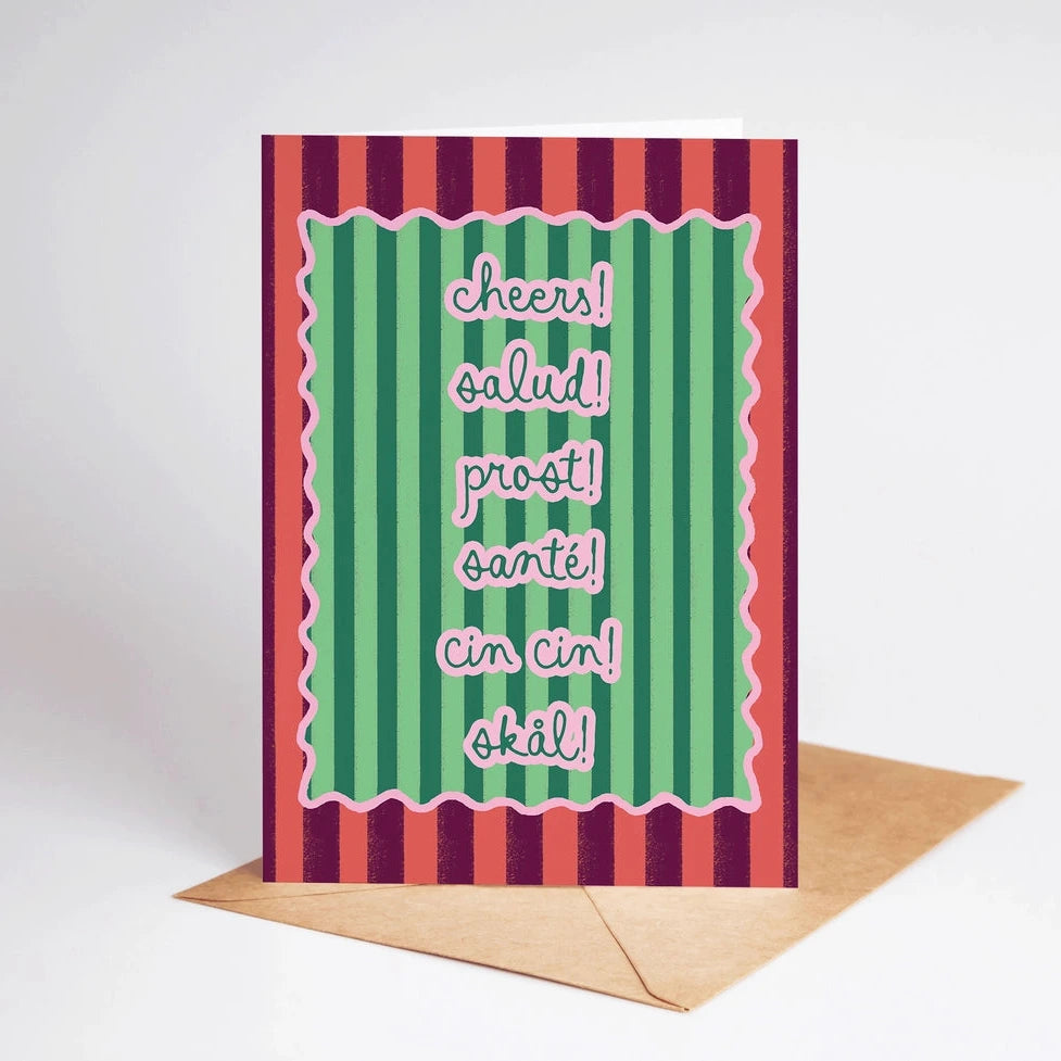 Greeting card featuring colorful striped design with the text "cheers! salud! prost! santé! cin cin! skål!" on a green background, accompanied by a brown kraft envelope, representing Brave Daughters' encouragement theme.