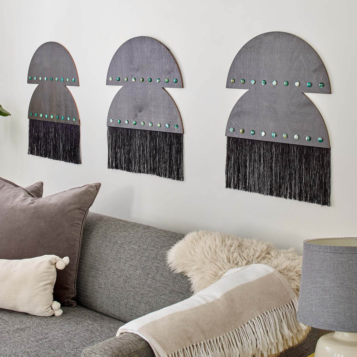 Half-moon shaped Ascension Wall Hanging in ebony stained wood with brass settings and agate cabochons, displayed on a wall above a gray sofa, featuring decorative fringe, emphasizing sustainable craftsmanship.