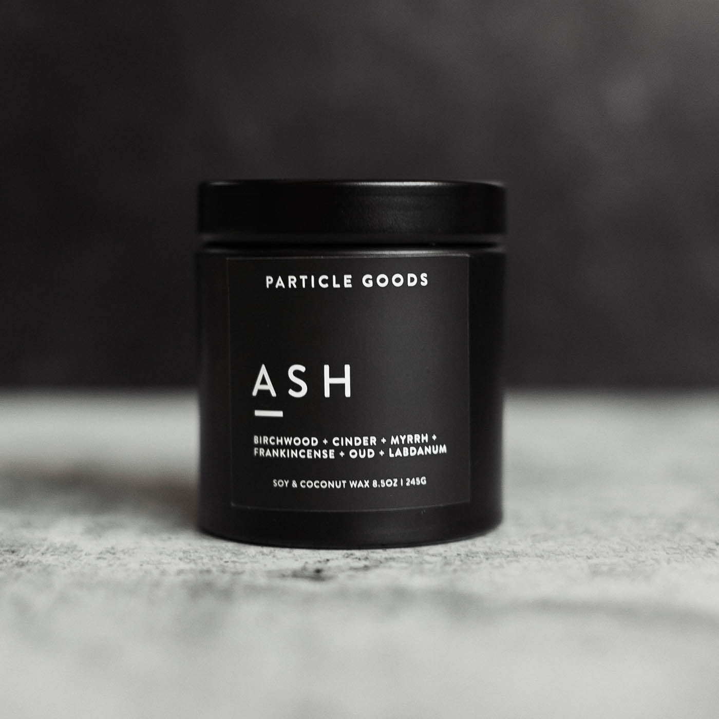 Candle labeled "ASH" by Particle Goods, featuring scent notes of birchwood, cinder, myrrh, frankincense, oud, and labdanum, in a sleek black jar on a textured surface.