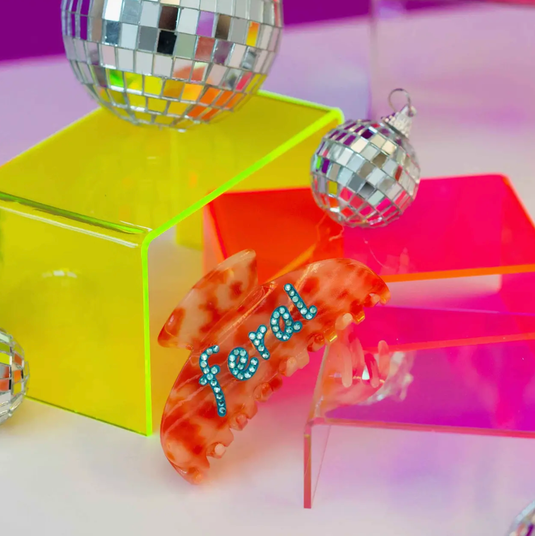 Feral Rhinestone Claw hair accessory in vibrant red and orange tortoise design with blue rhinestones, surrounded by colorful acrylic blocks and disco balls.