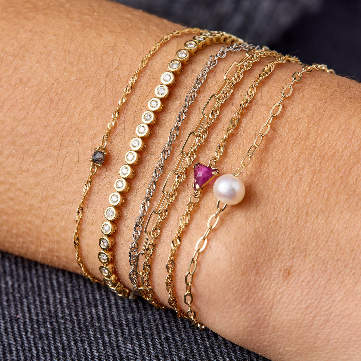 Custom permanent bracelet stack from Brave Daughters with 14k gold paperclip and helix chains, black diamond bezels, ruby, and pearl — welded fine jewelry for meaningful everyday wear.
