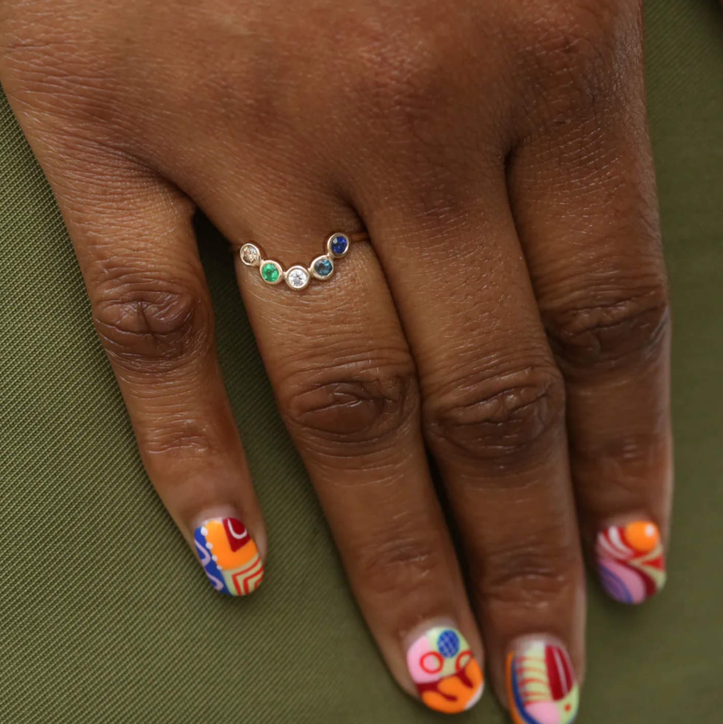 Hand with colorful nail art and a 14k gold ring with gemstones and diamonds on a green fabric background