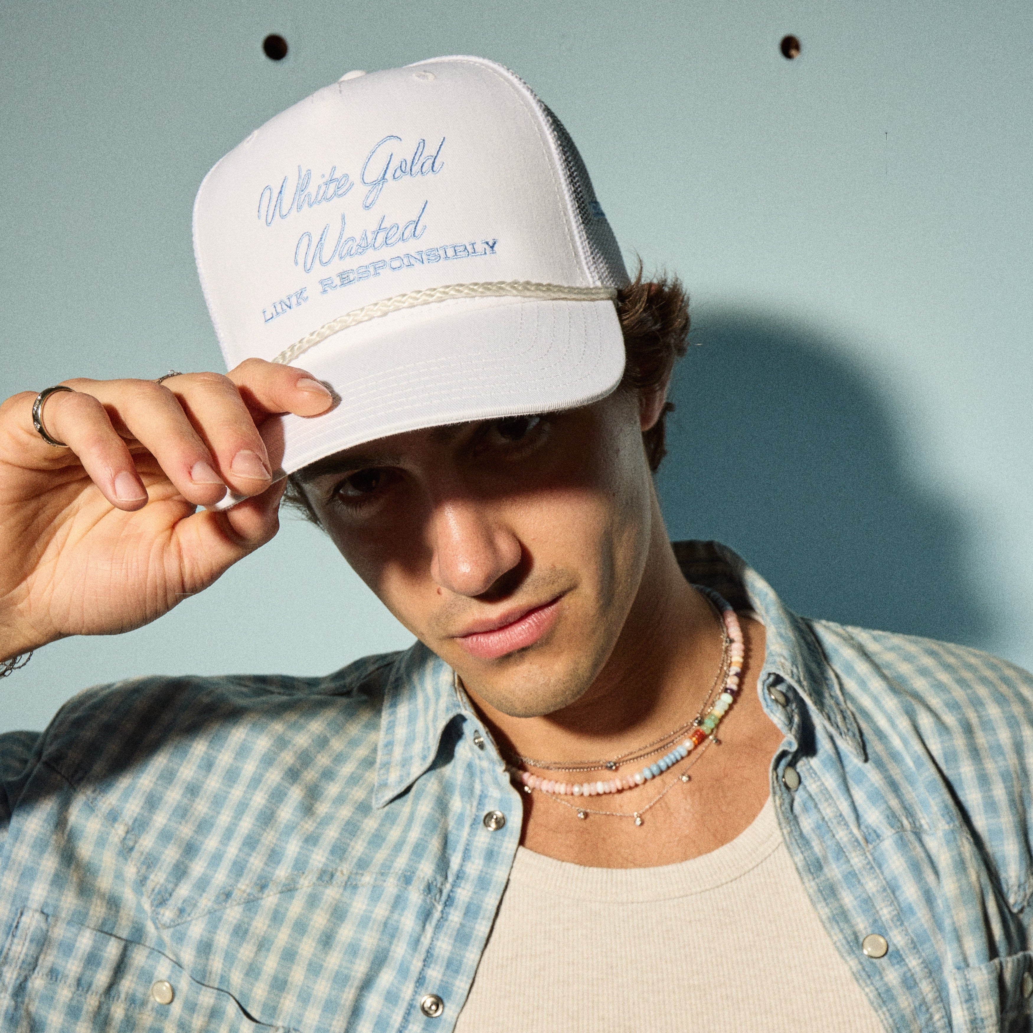 White Gold Wasted trucker hat worn by model, featuring embroidered text and casual summer attire, promoting Brave Daughters lifestyle products.