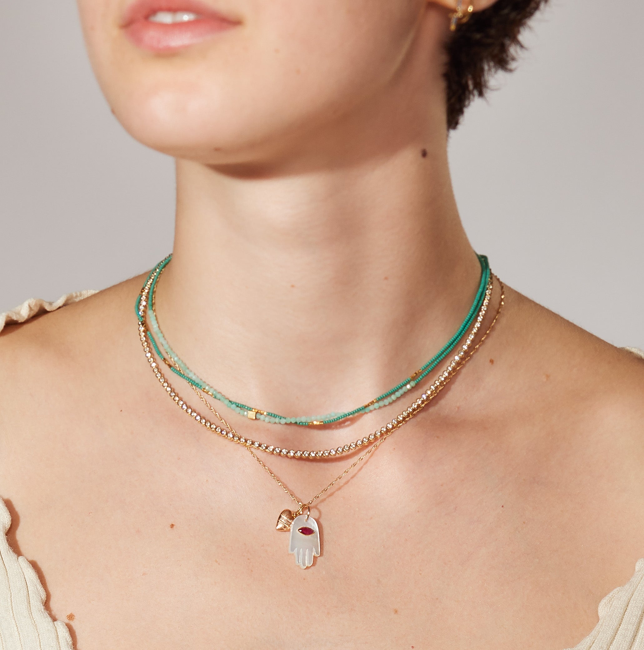 Layered necklaces featuring teal and gold beads, with a prominent hand-shaped pendant adorned with a red gem, worn by a model.