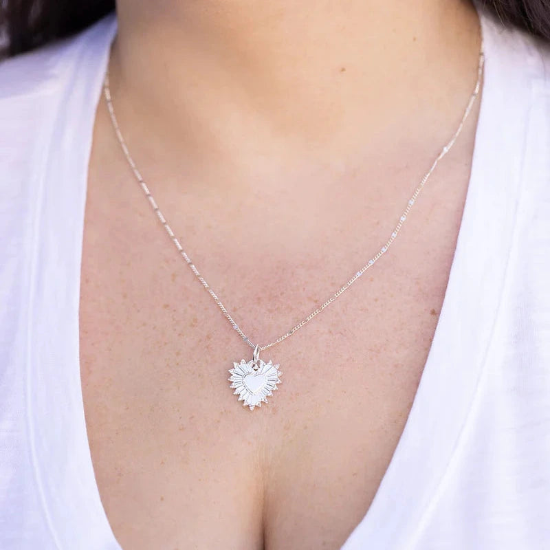 a silver heart charm on a silver chain on the neck of a person wearing a white v neck t-shirt