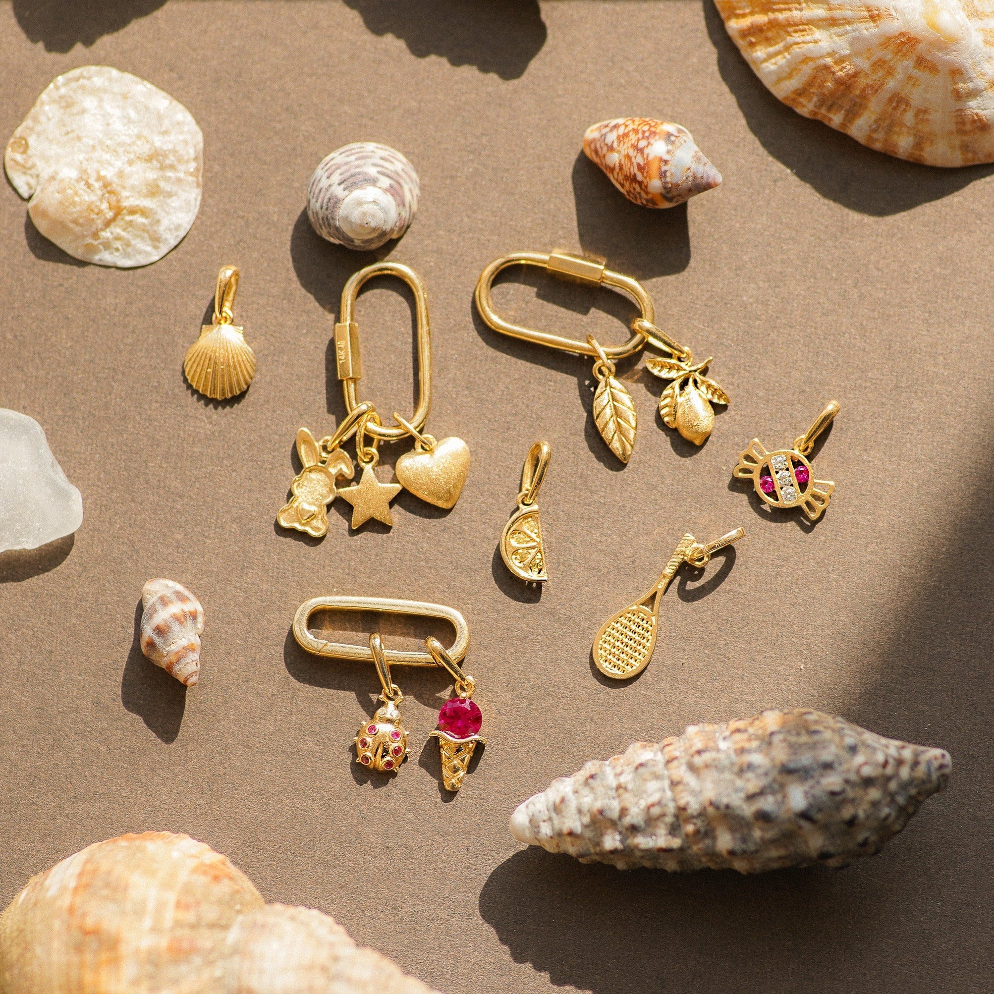 Gold jewelry charms with gemstones on a sandy surface with seashells.