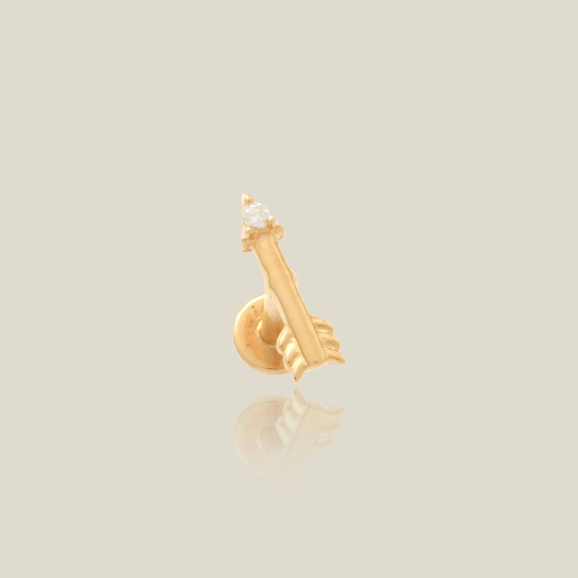 a gold stud earring in the shape of an arrow with a white prong set stone at the tip over a beige background