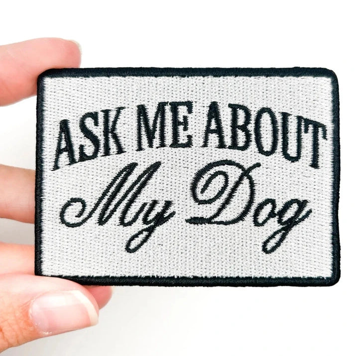 a black and white patch with text reading ask me about my dog held by a hand over a white background