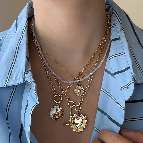 A close-up of a person wearing layered gold necklaces with distinct charms, including a yin-yang symbol, a heart with a clock face, and a sunburst design, styled over a light blue striped button-down shirt.