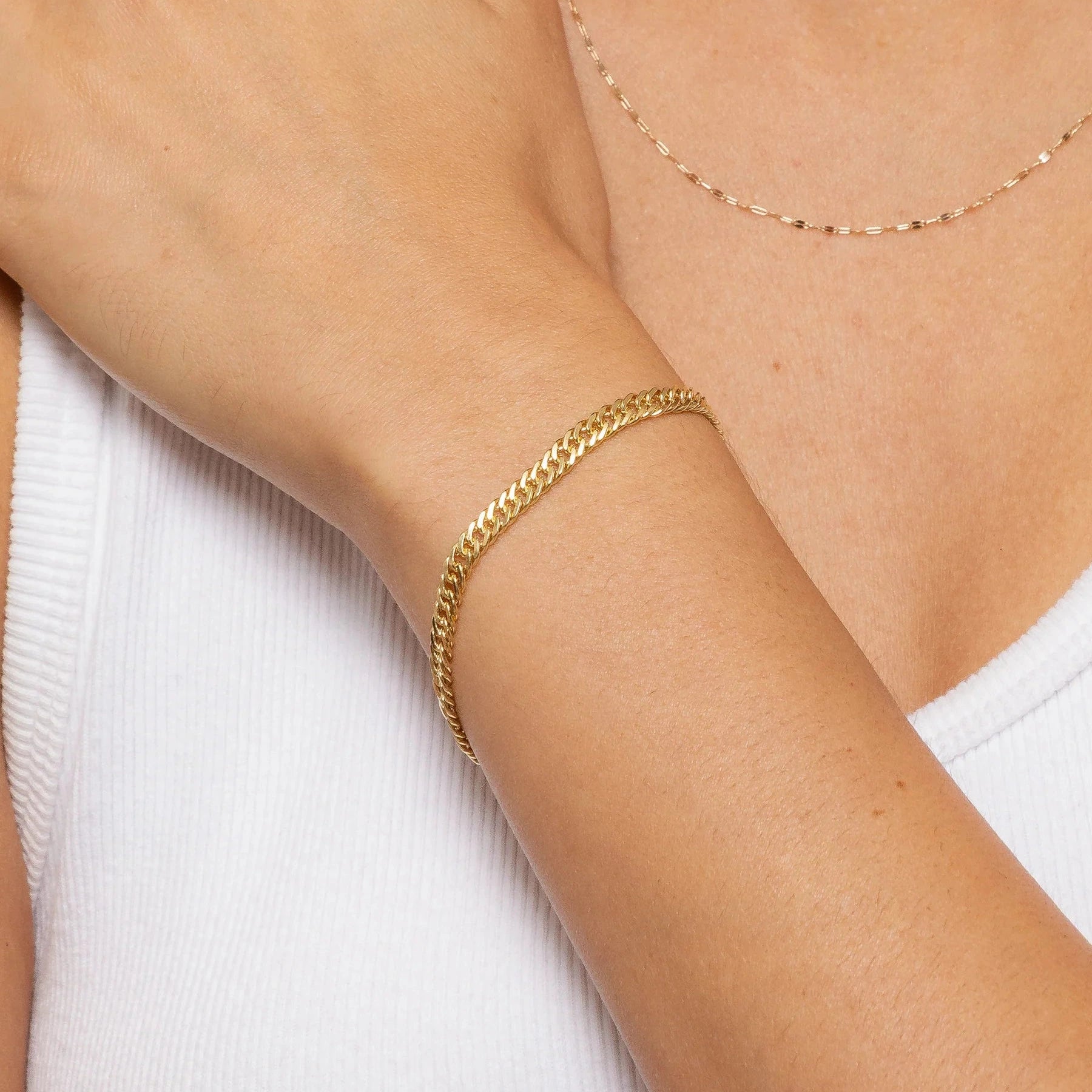A close-up of a person wearing a delicate gold chain bracelet on their wrist, paired with a matching fine gold chain necklace. The person is wearing a white ribbed tank top.