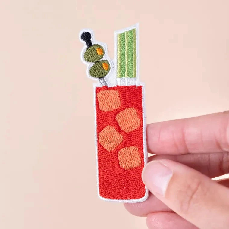 a red green and white iron on patch of a bloody mary with olive garnish, behind held by a hand over an off white background