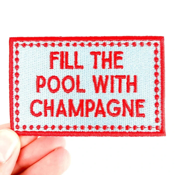 a red and blue iron on patch with texting reading fill the pool with champagne held by a hand over a white background