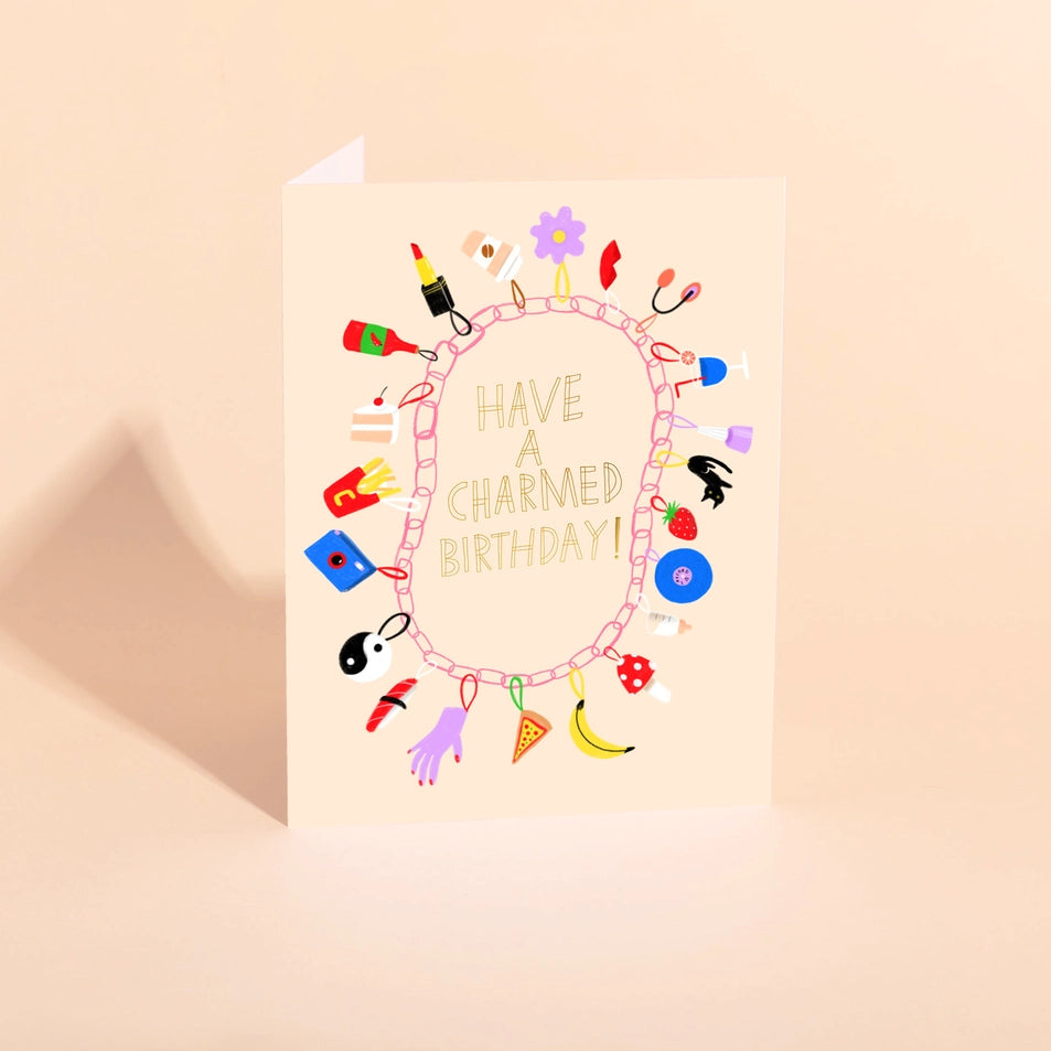 a peach colored greeting card with text reading have a charmed birthday surrounded by illustrations of charms on a bracelet, all over a neutral background