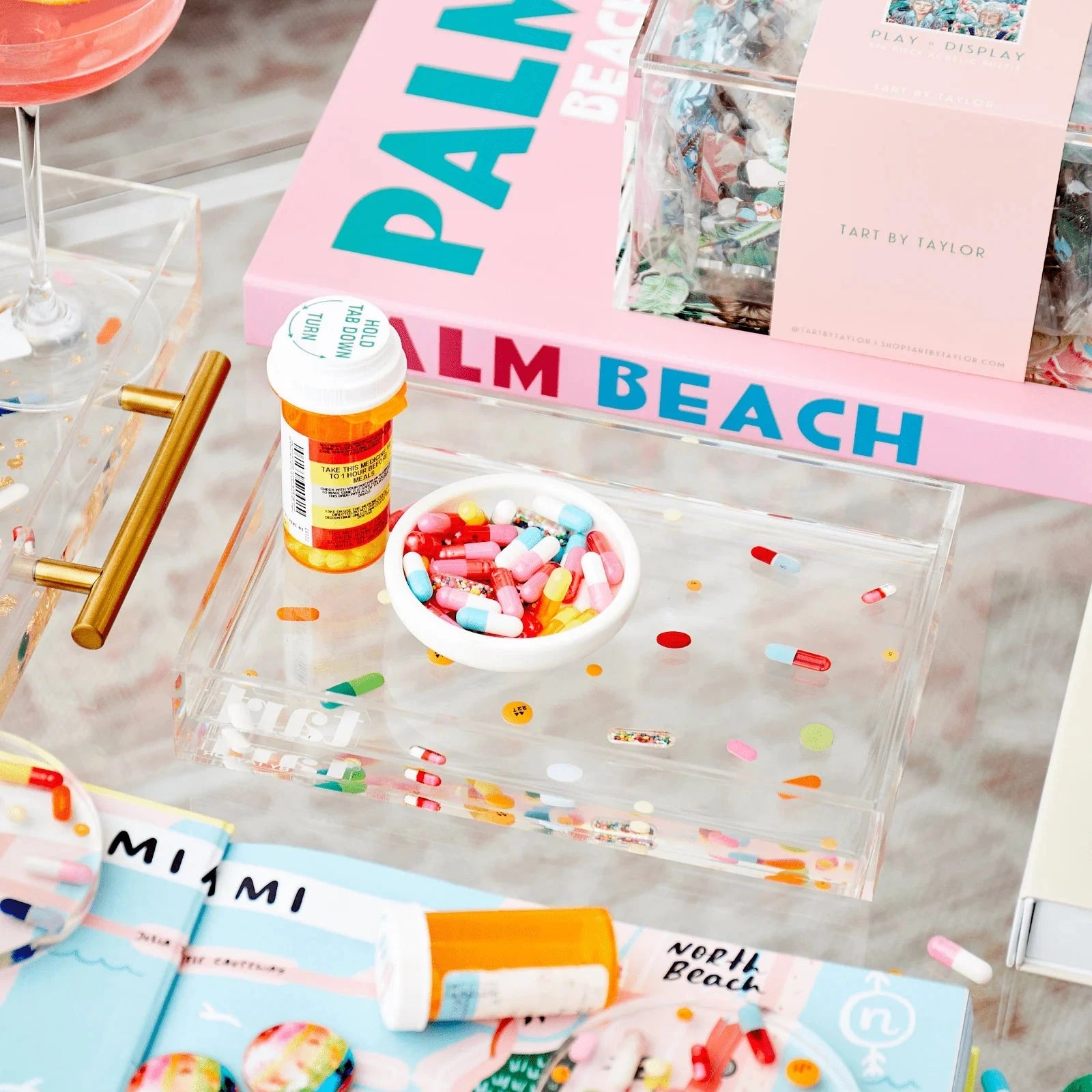 Decorative setting with 'Palm Beach' book, colorful candies, and a cocktail on a glass table.