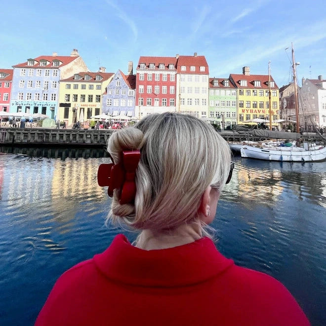 Person in a red coat looking at colorful buildings by a waterfront with a red bow claw clip
