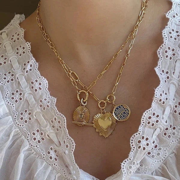 A close-up of a person wearing a layered gold necklace with multiple charms, including a heart-shaped pendant, a sunburst pendant, and a round striped charm, paired with a white eyelet lace blouse.