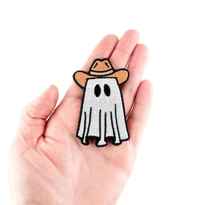a white black and tan iron on patch of a ghost wearing a cowboy hat being held by a hand over a white background