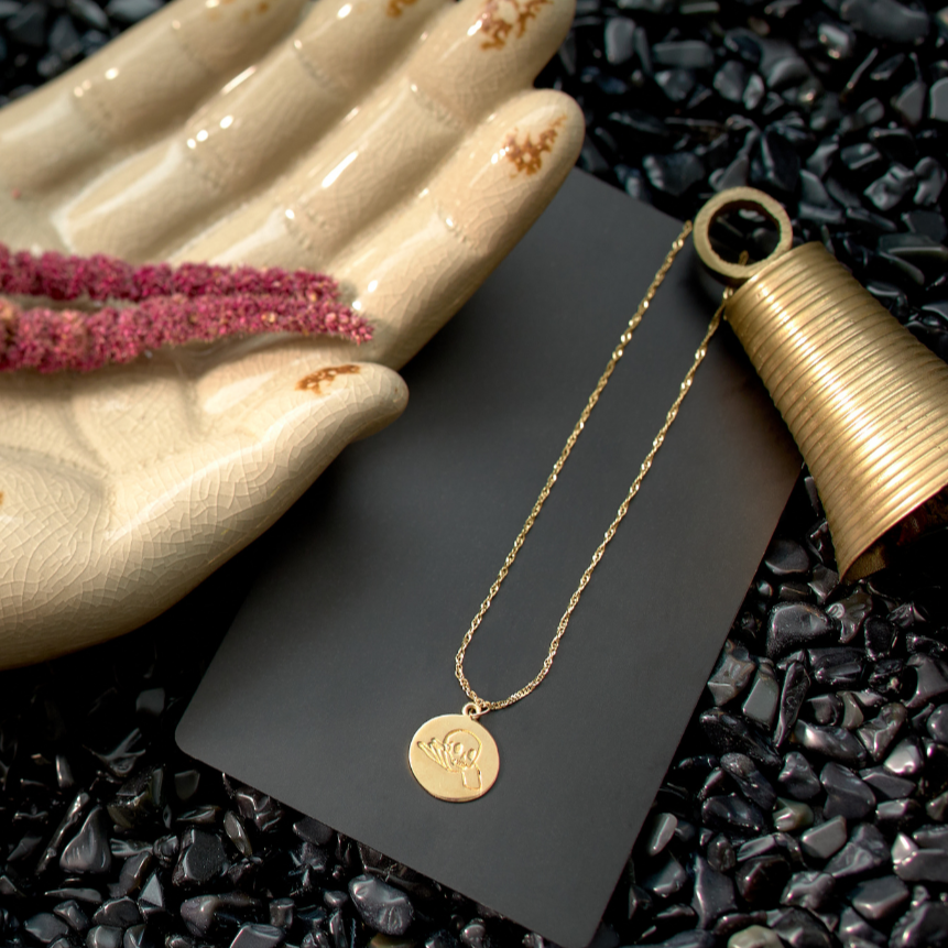 Gold necklace on a dark surface with a hand and pink flower in the background