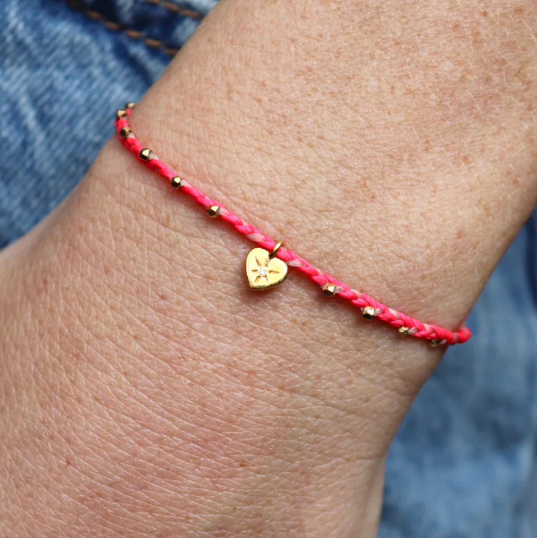 Pink nylon bracelet with a gold heart charm on a wrist against a denim background