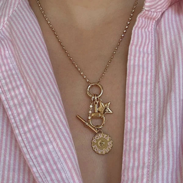 A gold charm necklace worn by a person in a pink and white striped button-down shirt. The necklace features a delicate gold chain, a top ring, and three charms: a small star, a miniature sword, and a round medallion charm with a central 'C' detail.