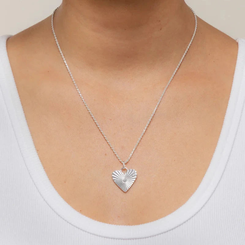 a silver heart shaped pendant charm on a necklace worn on the neck of a person wearing a white top