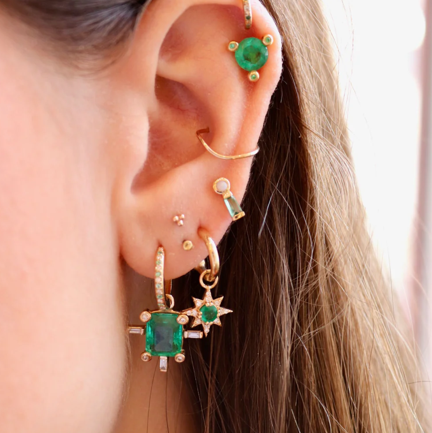 Close-up of an ear with multiple 14k gold and emerald green earrings.