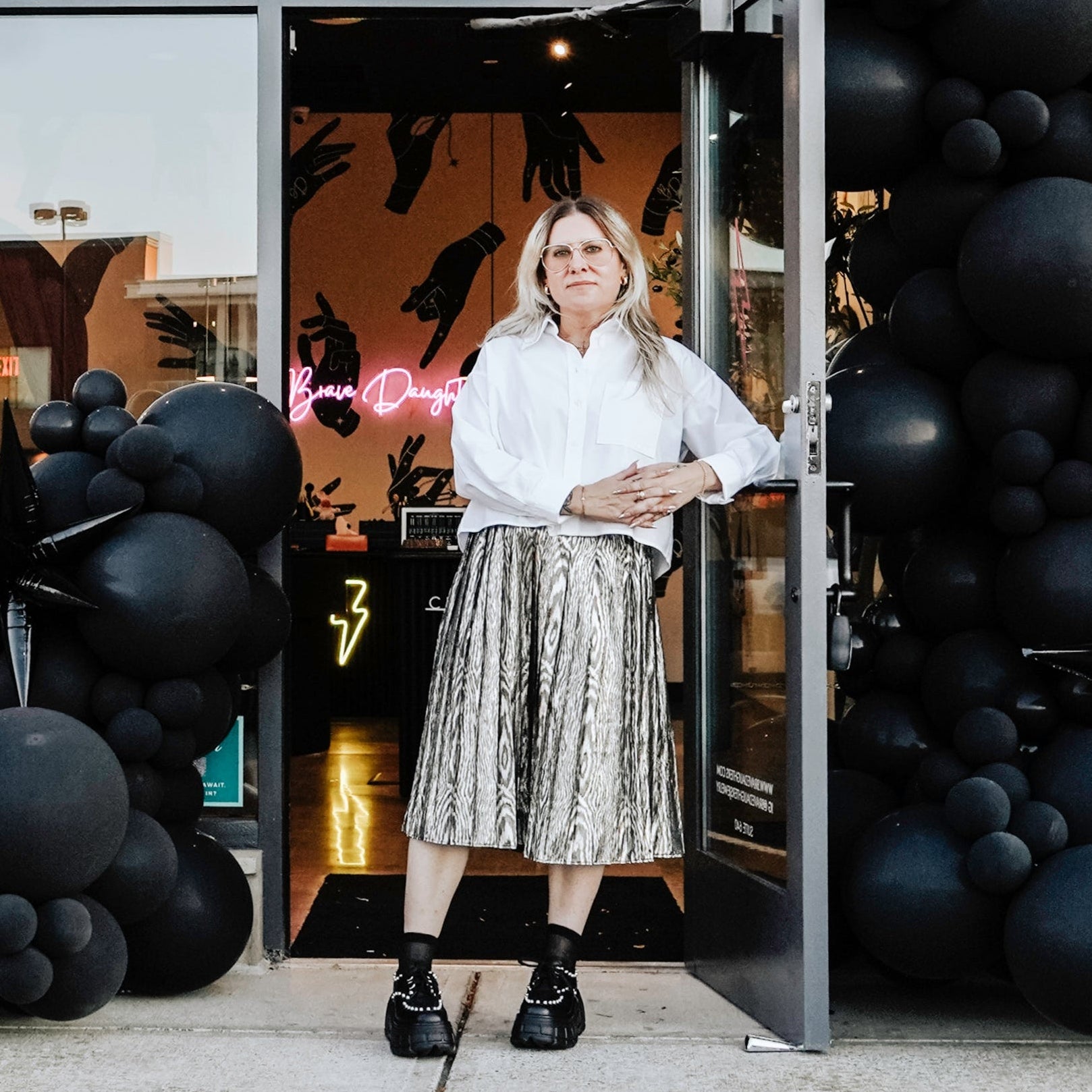 Woman standing in front of a store with 'Brave Daughters' sign and black balloons.