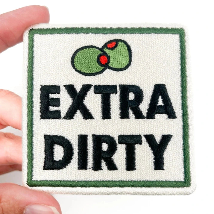 a cream black and green iron on patch with text reading extra dirty being held by a hand over a white background