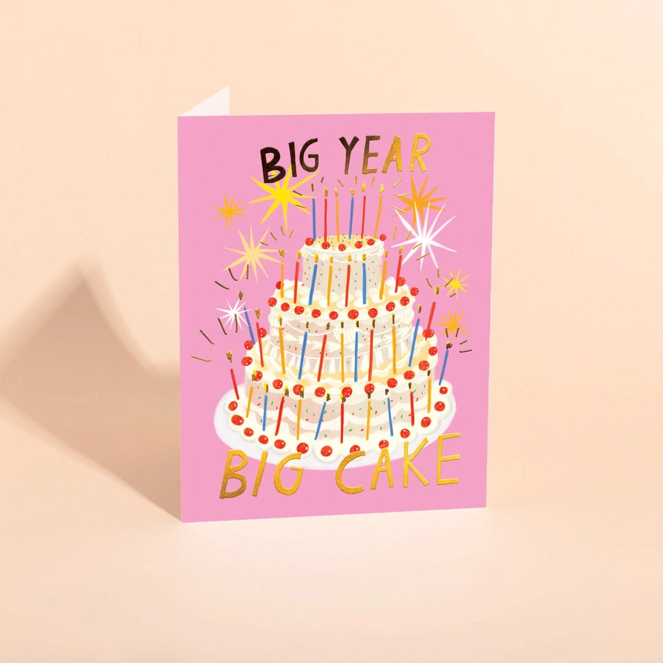 a greeting card with a pink background and an illustration of a three tiered birthday cake with dozens of candles and gold text reading big year, big cake