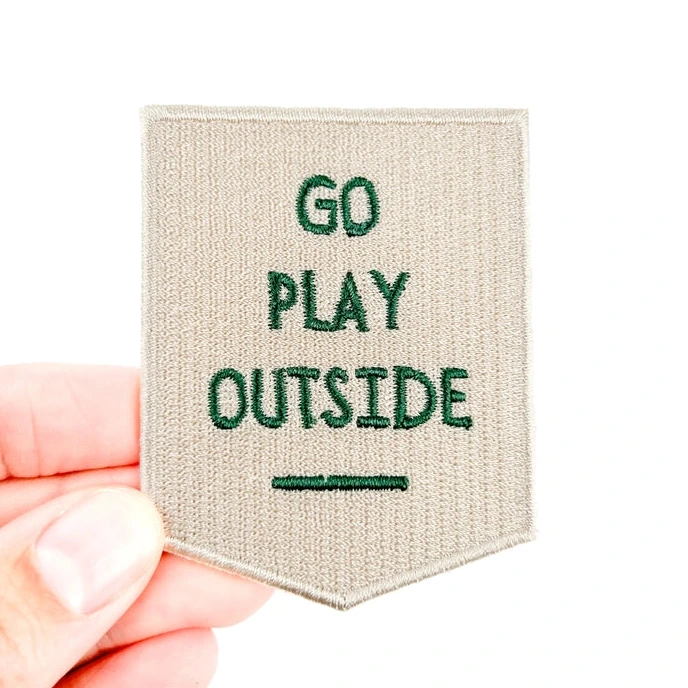 a khaki and green iron on patch with text that reads go play outside held by a hand over a white background