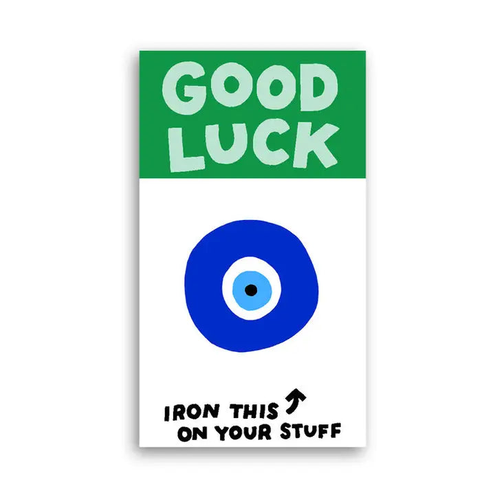A rectangular sticker or decal with a green top section reading 'GOOD LUCK' in white letters, a large blue evil eye symbol in the center, and black text at the bottom reading 'IRON THIS ON YOUR STUFF' with an arrow, shown on a white background.