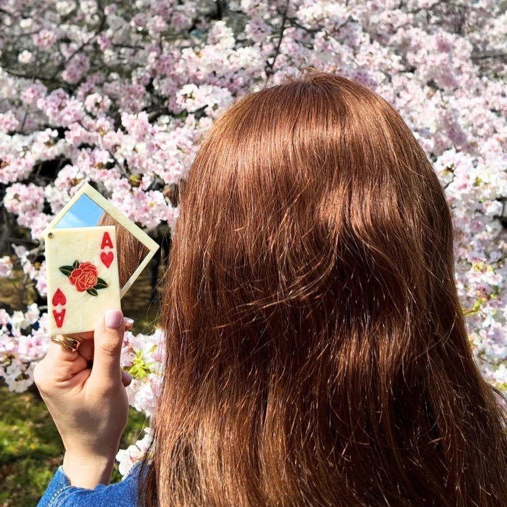 a person with red hair holding a white and red compact mirror in the shape of a playing card with the design of the ace of hearts and a floral motif with a cherry blossom tree background