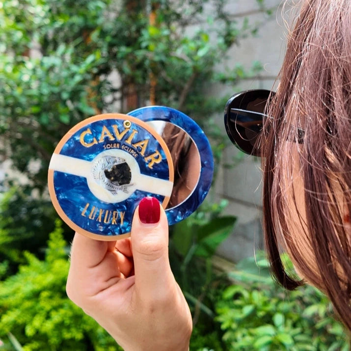 a person holding a blue and white compact mirror in the shape and with the detail of a tin of caviar with a natural verdant background