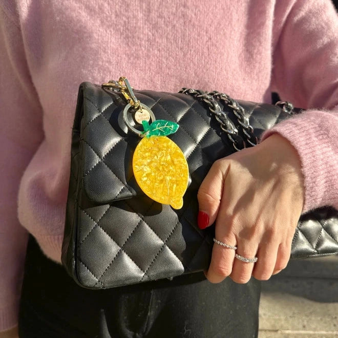 Person holding a black quilted handbag with a lemon charm, wearing a pink sweater.