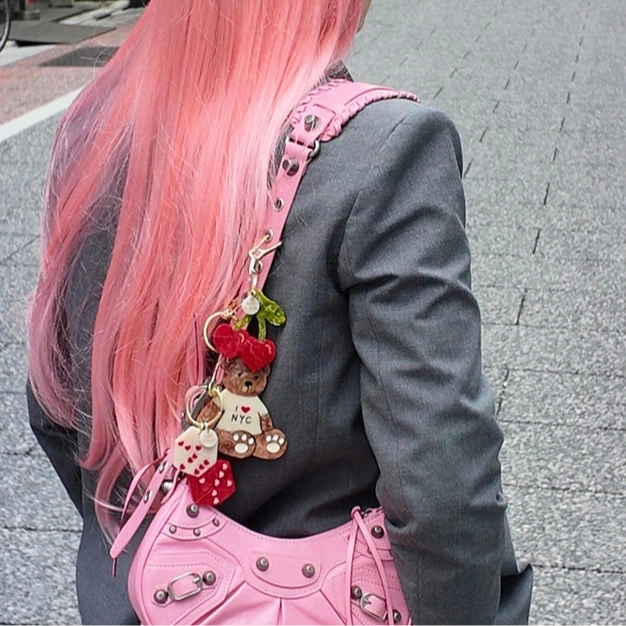 Person with pink hair and a black cap walking on a street with a dice keychain on her purse