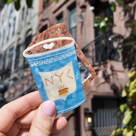 Hand holding a compact mirror in the shape of an nyc coffee cup with a cityscape background