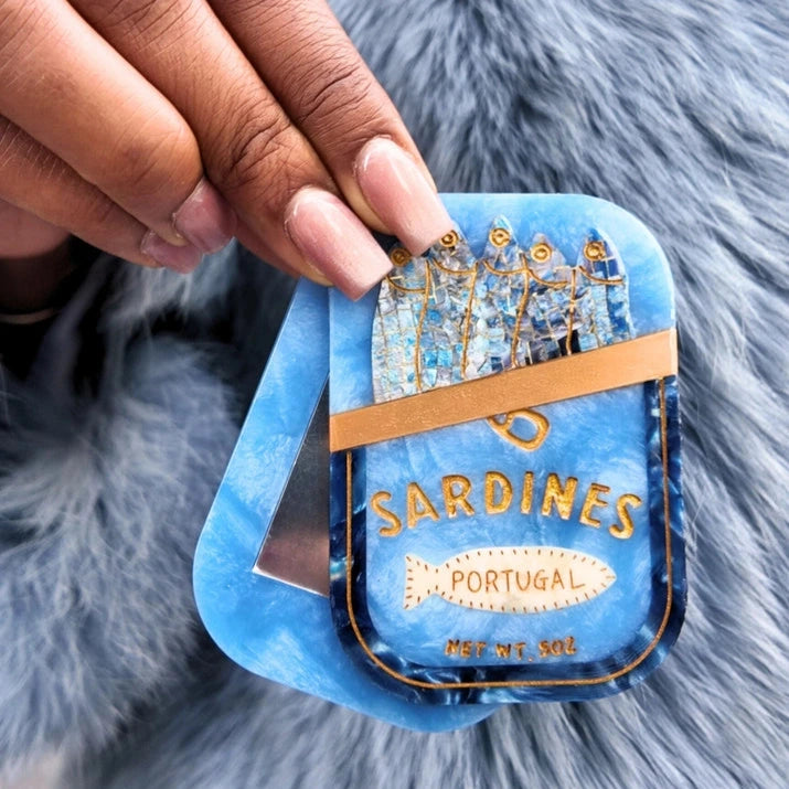 a hand holding a blue compact mirror in the shape and with the detail of a tin of sardines over a blue fuzzy coat background