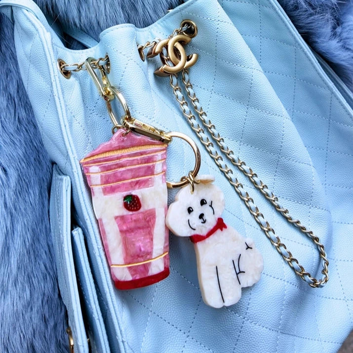 Close-up of a blue handbag with a pink smoothie cup and white dog keychain on a fluffy blue surface.