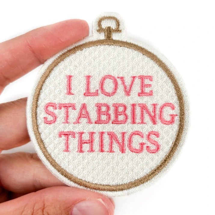 a white iron on patch with pink text reading i love stabbing things held by a hand over a white background