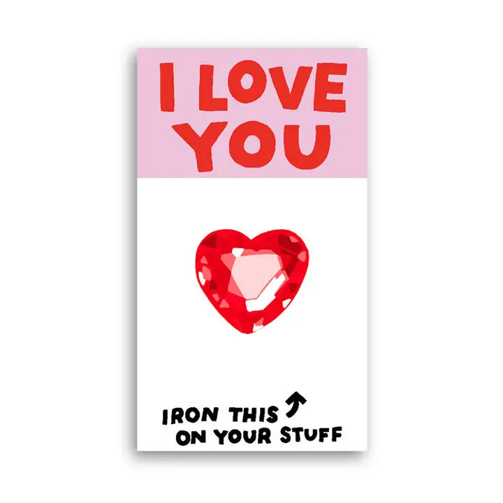 A rectangular novelty card or sticker with a pink top section reading 'I LOVE YOU' in red letters, a red faceted heart graphic in the center, and black text at the bottom that says 'IRON THIS ON YOUR STUFF' with an arrow pointing upward. The card is shown on a plain white background.