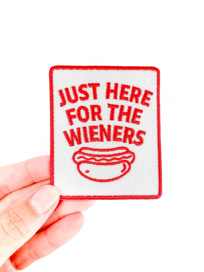 a red and white iron on patch with text reading just here for the wieners held by a hand over a white background
