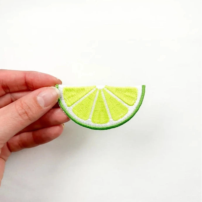 a green and white iron on patch of a slice of lime held by a hand over a white background