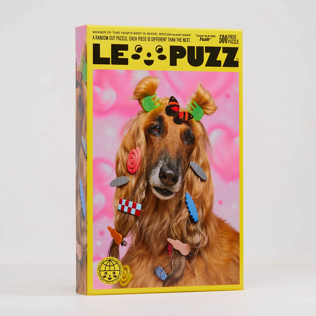 A boxed jigsaw puzzle titled "LE PUZZ" standing upright on a light gray studio background. The front of the box features a brown dog with colorful hair accessories against a bright pink background.