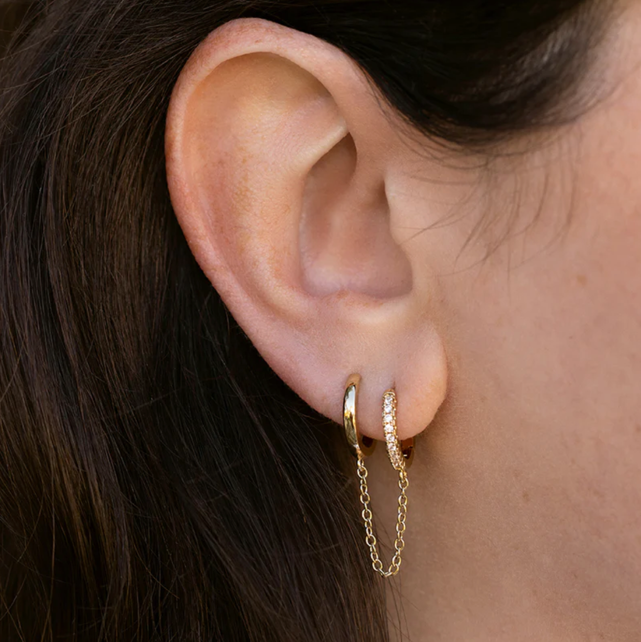 Close-up of an ear wearing gold hoop earrings with a chain detail.