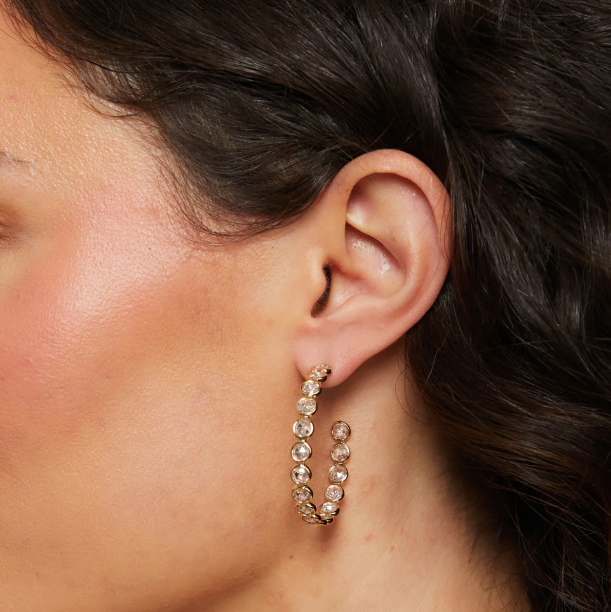 Close-up of an ear wearing a gold hoop earring with clear stones.