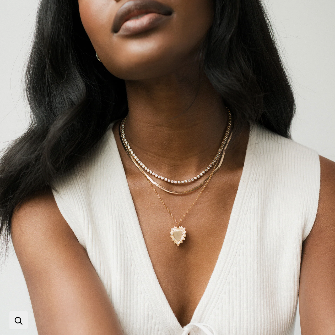Woman wearing a white top and gold necklaces with a heart pendant on a plain background