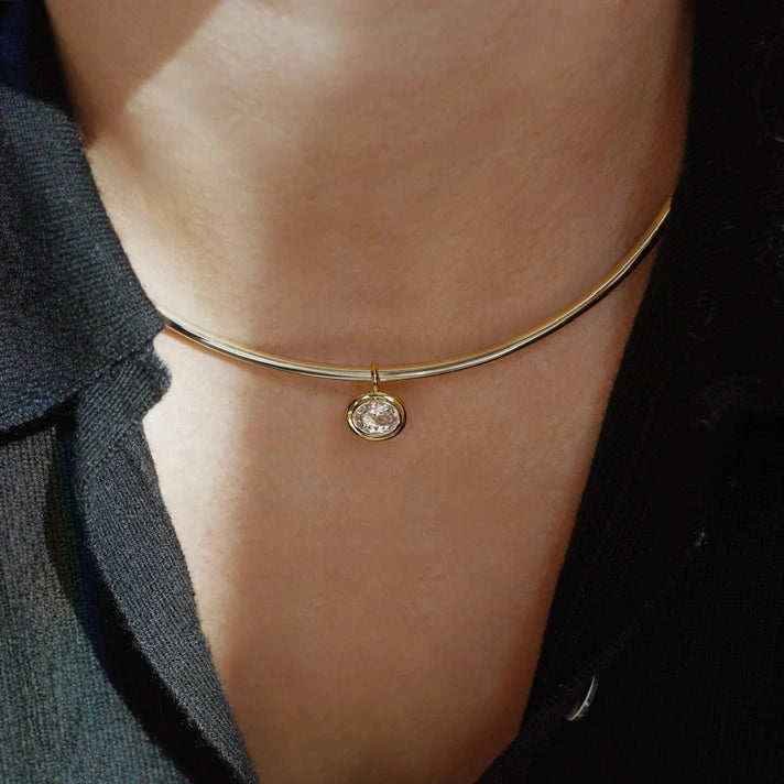 an oval gold charm with a bezel set white sapphire on a gold chain worn with a black collared shirt