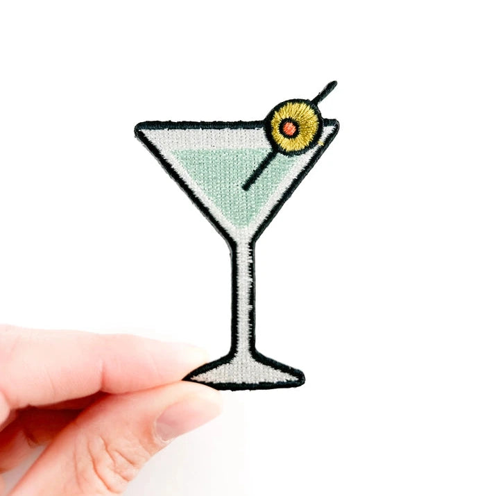 an iron on patch in the shape of a martini glass with an olive garnish held by a hand over a white background