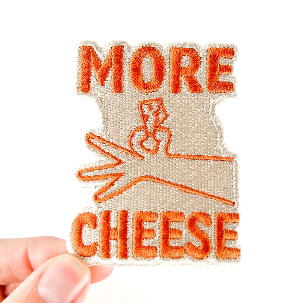 a brown and orange patch with text reading more cheese and hand illustration with a white background