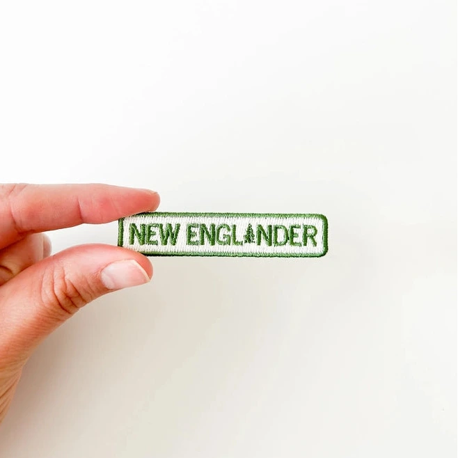a green and white iron on patch with text reading new englander held by a hand over a white background